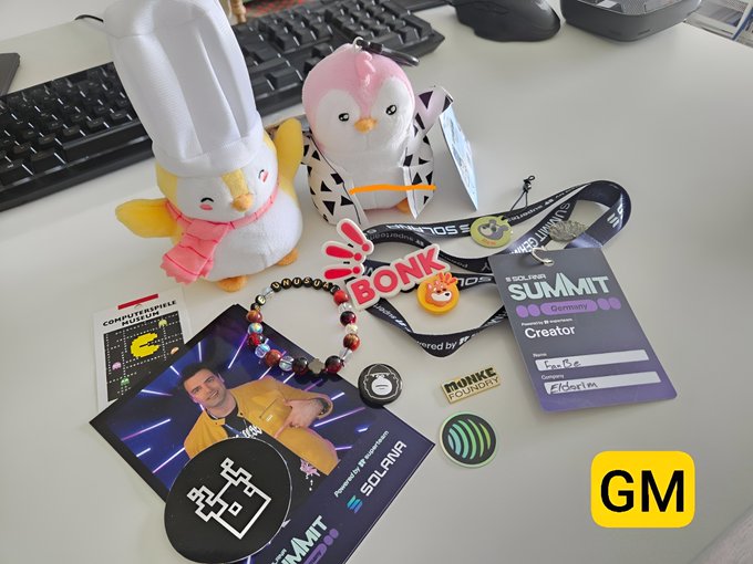 The image features a desk with various items scattered across its surface, including a keyboard, a mouse, a stuffed penguin toy, a badge, and several other small objects such as stickers and magnets. The main focus of the image is a post on social media that reads "GM Some memories are just unforgettable, like the Solana Summit Germany". This post seems to be related to the desk items in the foreground, possibly indicating an event or gathering where these items were used for taking photos or sharing experiences with friends and followers.