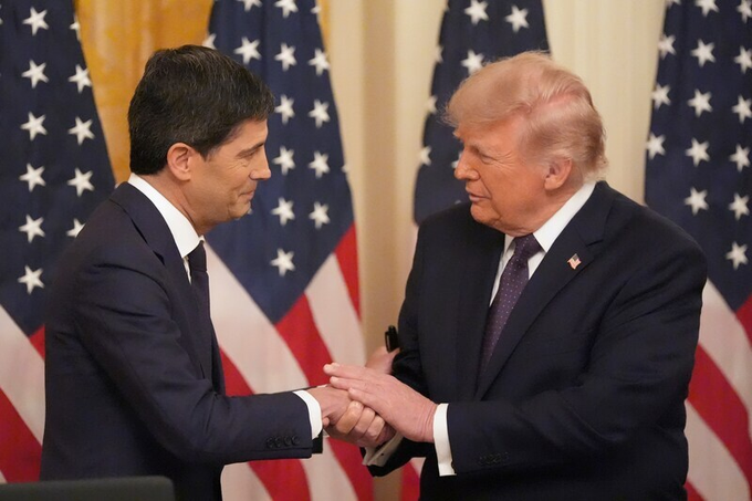 Two men in dark suits shaking hands in front of American flags.