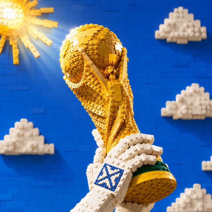A LEGO recreation of the FIFA World Cup trophy held up against a blue sky with white brick clouds and a golden sun, with a LEGO hand wearing a Scotland flag wristband.