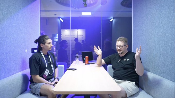 Two people seated at a table in a blue-lit room during what appears to be a podcast interview; the man on the right is gesturing with his hands while speaking.