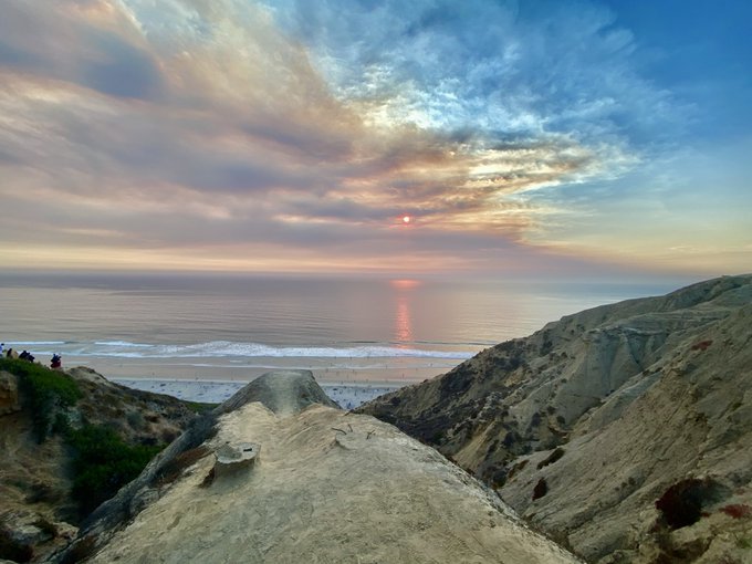 A scenic sunset view over a coastal landscape, featuring dramatic clouds, a calm ocean with gentle waves, and rugged sandy cliffs in the foreground where a few small figures can be seen.