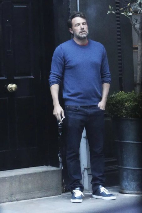 A man in a blue sweater, dark jeans, and sneakers standing outside a building smoking a cigarette, photographed in a candid style.