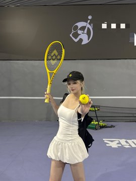 A young woman in a white tennis dress and black cap poses on a blue court, holding up a yellow tennis racket and a yellow "Binance" branded tennis ball, appearing energetic before her workout as referenced in the post.