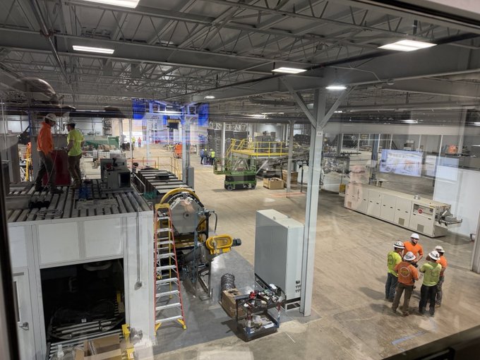 An industrial manufacturing plant or factory floor with workers in safety gear, a yellow scissor lift, conveyor systems, and various machinery under bright overhead lighting.