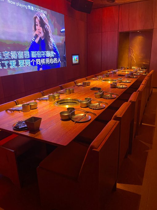 A dimly lit, reddish private dining room with a long wooden table set with grills and plates, a row of tan leather chairs, and a karaoke screen displaying a singer and Chinese lyrics on the wall.