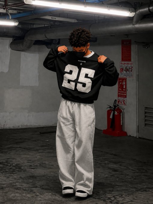 A person in a parking garage wearing an oversized black sweatshirt with a large white '25' and light gray baggy sweatpants, holding the collar of the sweatshirt.