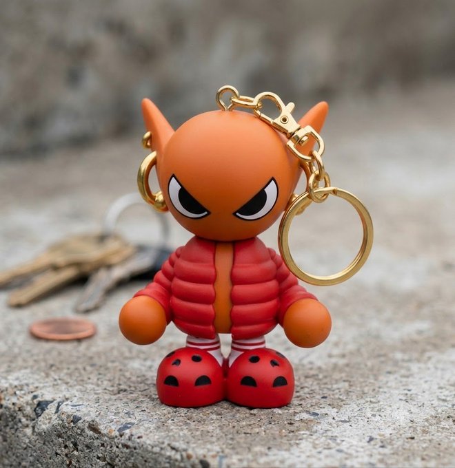 A small orange figurine keychain with an angry expression, wearing a red puffer jacket and red shoes, attached to keys resting on the pavement.