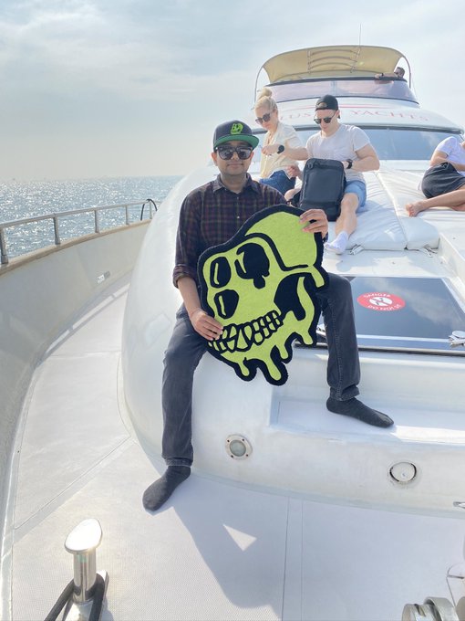 A man in a green cap, sunglasses, and plaid shirt sits on the bow of a yacht, proudly holding up a large neon-green Bored Ape skull rug. Behind him, other members relax on cushioned seats near a "Bored Ape Yacht Club" banner, capturing the luxurious, community-driven spirit of the famous NFT collection.