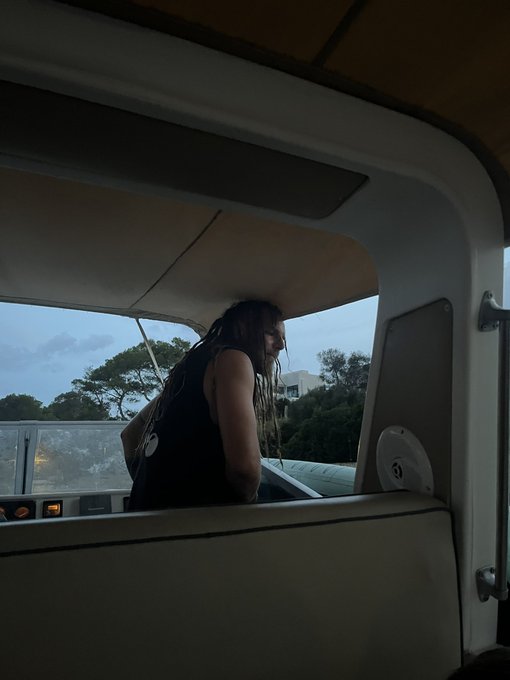 A silhouetted figure with long dreadlocks looking out the window of a bus or train at dusk, with a lanyard visible around their neck.