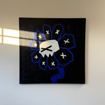 A piece of canvas art featuring a white skull surrounded by a blue flower-like shape with 'X' marks on the petals, hanging on a white wall and lit by sunlight.