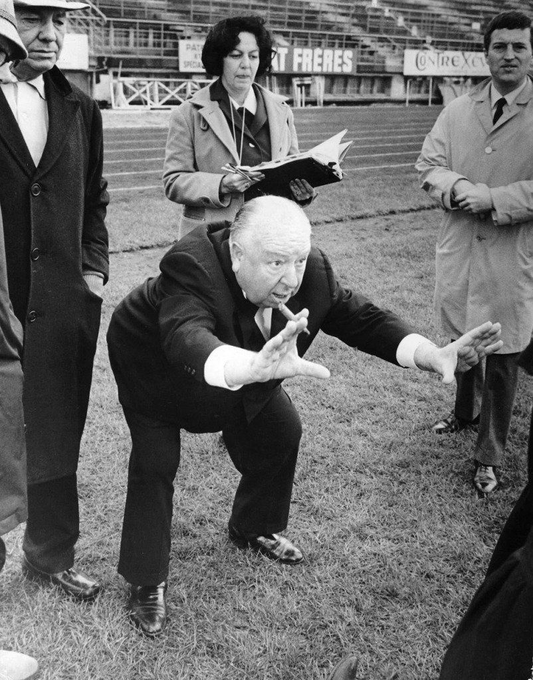A black-and-white photo shows a man in a suit crouched on a sports field, gesturing with both arms, while a woman with a notebook and two men in coats and hats observe.