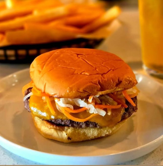 Close-up of a cheeseburger with shredded carrots, onions, and melted cheese on a white plate, with fries and a glass of orange beverage in the background.