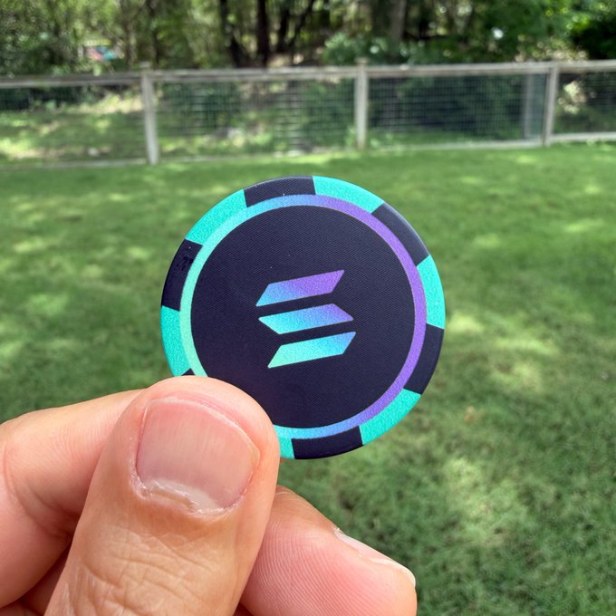 The image features a hand holding up a small, blue and green poker chip with the Solana logo on it. The background of the photo shows a grassy area with trees visible behind the person holding the chip. The text "Solana Poker Season" is also present in the image. This suggests that the Solana cryptocurrency has recently gained popularity or is associated with the poker season, possibly indicating an increase in interest in both Solana and poker-related activities during this time of year.