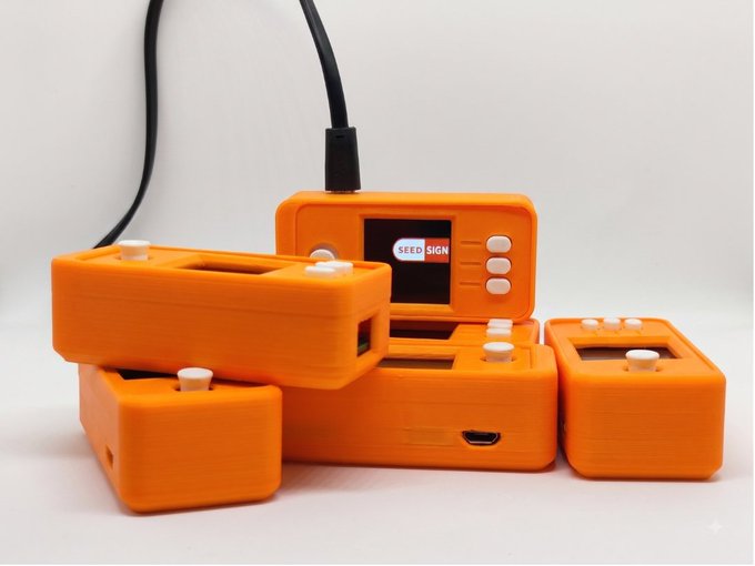 A collection of orange 3D-printed SeedSign hardware wallet devices, with the central unit displaying a screen and connected via a USB cable.