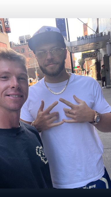 A selfie photo of two men on a city street; the younger man on the left is smiling while the bearded man on the right wears a white t-shirt, baseball cap, sunglasses, and a chain necklace, holding up hand gestures.