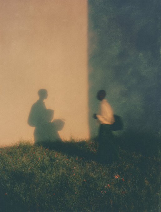 An artistic photograph showing two figures—one as a sharp silhouette against a warm beige wall, the other blurred in motion wearing white—standing in a grassy field at dusk.