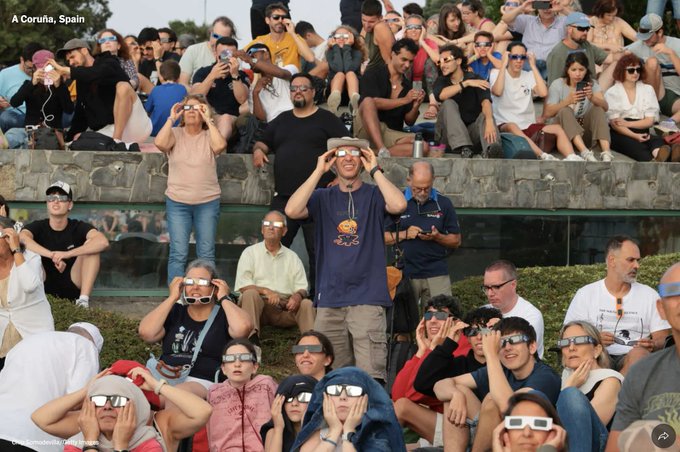 A photograph labeled 'A Coruña, Spain' captures a crowd of spectators outdoors, all wearing protective solar eclipse glasses and looking upward to view an astronomical event.