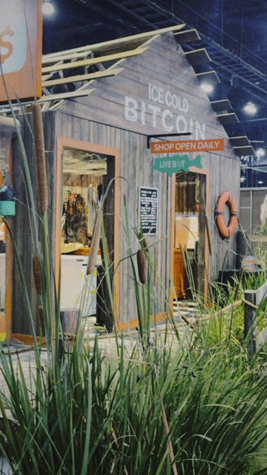 A quirky trade show booth designed like a rustic beach shack displays the words "ICE COLD BITCOIN" and "SHOP OPEN DAILY LIVE BIT" on its wooden facade, complete with a life preserver and cattail reeds in the foreground, fitting the playful branding mentioned by Complex.