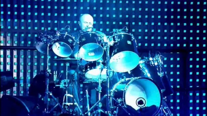 The image features a man playing drums, with his drum set positioned in front of him. He appears to be wearing headphones while performing. The background consists of blue lights that create an interesting visual effect as they illuminate the drummer's performance area. There is also text present in the image, which reads "mood. $TIBBIR". This suggests that the image might have been taken during a live music event or concert where the mood and atmosphere are important aspects to consider for the audience.