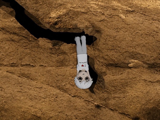 A digitally manipulated image of an anime-style character wearing a t-shirt that says 'I ♥ EXPLOSION' hanging upside-down from a crevice in a rocky, brown cliff face.