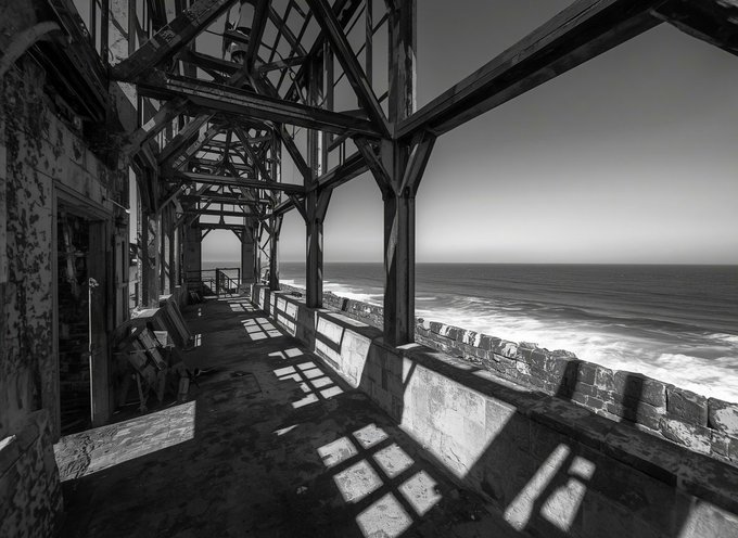 A black and white photograph looking down a corridor of an abandoned building with exposed wooden beams, where sunlight casts geometric shadows on the floor and a stone wall overlooks the ocean waves.