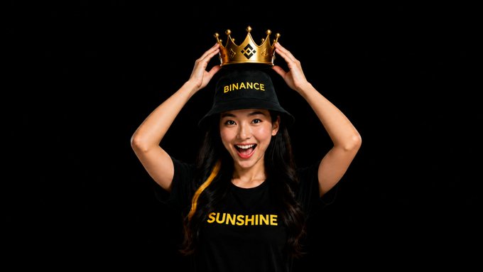 A smiling woman wears a black t-shirt with 'SUNSHINE' in yellow text and a black bucket hat with 'BINANCE' in yellow text, while holding a golden crown on her head.
