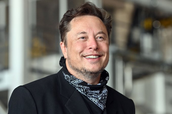 The image shows Elon Musk smiling broadly, dressed in a dark blazer with a black and white patterned scarf, appearing in what looks like an industrial or warehouse setting. The image pairs with the post's quote about Musk's perspective on money as an information system, visually representing him as the speaker behind these thoughts on the future of Bitcoin, crypto, and frictionless financial systems.