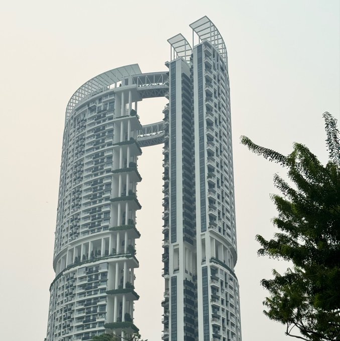 A towering skyscraper with twin towers connected by sky bridges and topped with curved crown-like structures stands against a hazy sky, with tree foliage in the foreground. The image relates to the post by showcasing an iconic piece of Singapore's modern architecture, likely one of the city's distinctive residential towers, serving as a scenic backdrop for the Token2049 event.