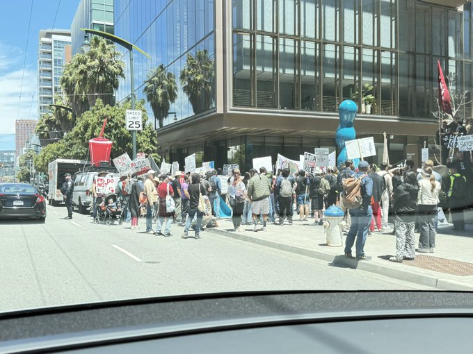 A crowd of protesters holding signs gathers on a city street outside a glass-fronted building, captured through a car windshield with a visible "SPEED LIMIT 25" sign in the foreground. Signs reading "STOP" and references to profit suggest demonstrators are rallying outside OpenAI's office, linking concerns about AI-driven inequality to the post's claim that artificial intelligence will produce new forms of communism amid rising productivity disparities.