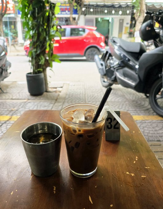 A glass of iced coffee with a black straw and a small metal cup sit on a wooden table outside a café, with motorcycles and a red car parked in the background.