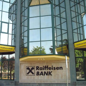 The image shows the exterior of a Raiffeisen BANK building with its distinctive logo and signage, reflecting the bank's presence referenced in the post about Raiffeisen Bank International's partnership with Bitpanda to offer crypto trading to its 18 million customers across 11 European markets.