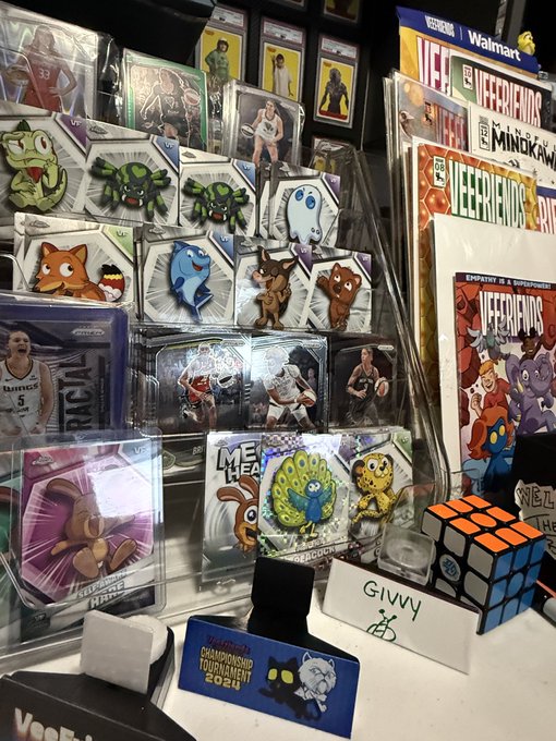 A display of merchandise, including Pokémon, NBA, and SpongeBob stickers, Veefriends cards, and a 2024 Champions League tournament pin, sits on a counter next to a 'GIVWY' cash sign.