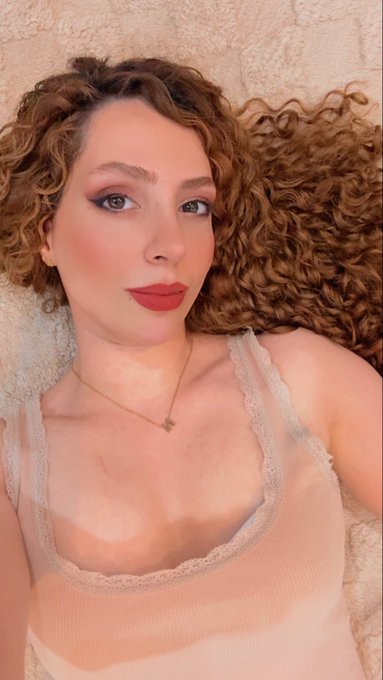 A selfie-style photo of a young woman with curly auburn hair, dramatic eye makeup, and red lipstick, wearing a peach-colored lace-trimmed top and a delicate gold necklace while lying on a beige textured surface.