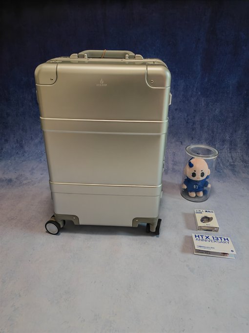A champagne-colored hard-shell suitcase standing next to a small plush mascot figure inside a clear cup and some small boxes on a blue background.