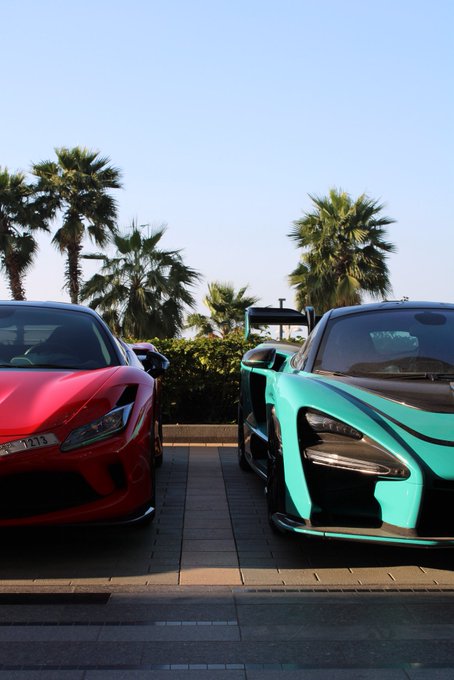 A red Ferrari and a teal McLaren are parked side by side on a brick driveway framed by palm trees. The "GM, if you say it back" caption plays on the rivalry between the two performance-car brands, suggesting that a Ferrari owner might reciprocate a McLaren driver's nod of mutual respect on the road.