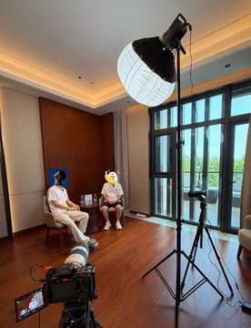 Two people are seated on chairs in a well-lit room with large windows while a professional camera on a tripod and a softbox light are set up to film them, with both faces blurred.