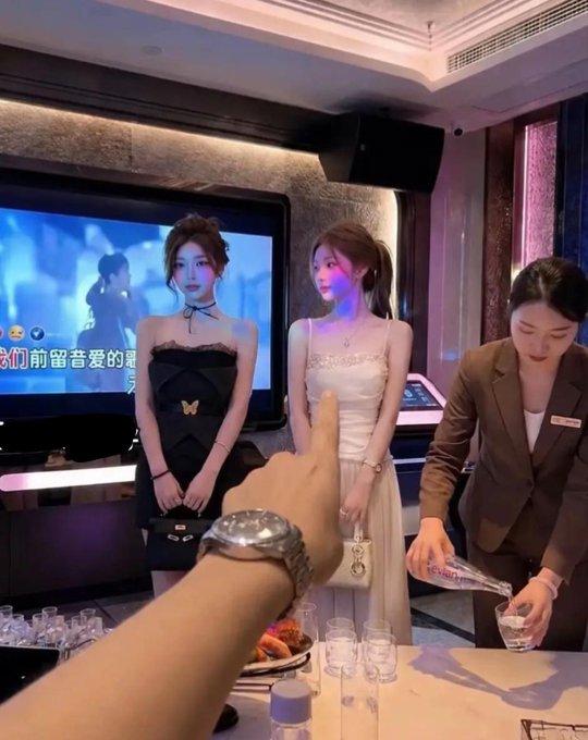 A man flashes his expensive watch toward two elegantly dressed women and a server at what appears to be a high-end KTV lounge, with Chinese text visible on the TV screen reading "我们曾亲爱的夏..."—capturing the smug swagger of someone celebrating a hypothetical 100x memecoin pump.