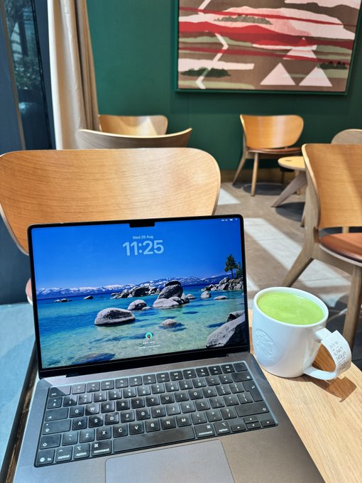 An open laptop sits on a wooden café table next to a Starbucks matcha latte, with the screen displaying a scenic lake wallpaper and the time 11:25.