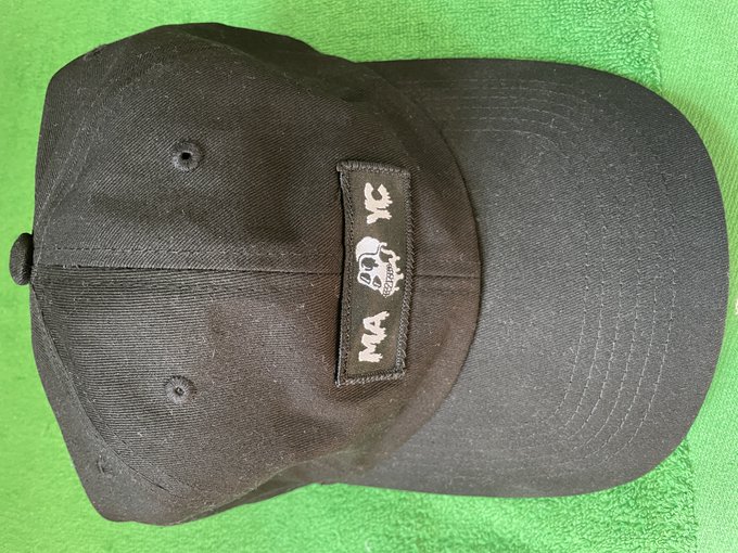 The image features a black baseball cap with a white skull patch on it. The hat appears to be new and unused, as indicated by its clean condition. The hat has a flat brim that covers most of the face, making it suitable for various outdoor activities or casual settings. The hat is placed on a green towel, which adds a pop of color to the scene. There are no other objects in the image besides the hat and the towel.