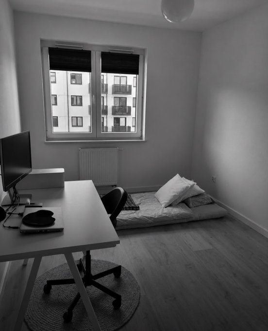The image shows a sparse, minimalist bedroom with a simple mattress on the floor, a basic desk with a monitor, and a chair in a modest room with large windows. The understated, bare-bones living space contrasts sharply with the extravagant claim of "$300,000 a month" in the caption, suggesting either ironic commentary or a stark depiction of how wealth doesn't necessarily translate to a lavish lifestyle in this particular space.