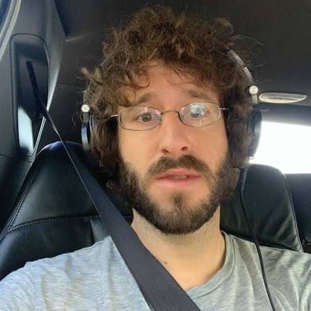A selfie of a bearded man with curly hair and glasses wearing headphones inside a car.
