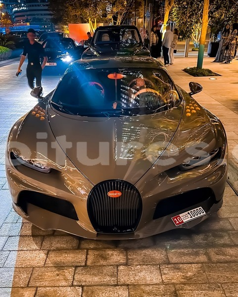 A gray Bugatti hypercar parked at night with headlights on, featuring a 'Dubai 10000' license plate and people standing nearby in a plaza setting.