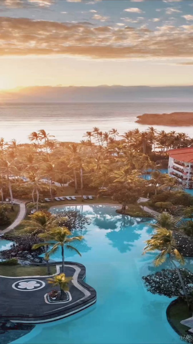 The image shows a beautiful tropical resort with a large pool surrounded by palm trees. The sun is setting behind the ocean, creating a warm glow over the scene. In the background, there are buildings that add to the overall ambiance of the location. The photo appears to be taken from an aerial perspective, providing a comprehensive view of the resort and its surroundings.