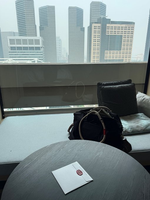 A view from inside a high-rise building looking out at city skyscrapers, with a backpack, pillow, and a sealed envelope on a round table in the foreground.
