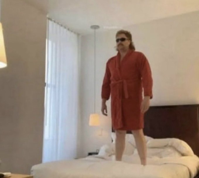 A man with long hair, a prominent mustache, and sunglasses stands on a bed wearing a red bathrobe in a modern bedroom setting.