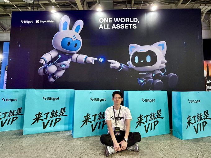 A person sits cross-legged in front of a large Bitget crypto exchange booth at a convention, featuring mascot characters and the slogan 'ONE WORLD, ALL ASSETS' with '来了就是VIP' (Once you're here, you're VIP) banners.