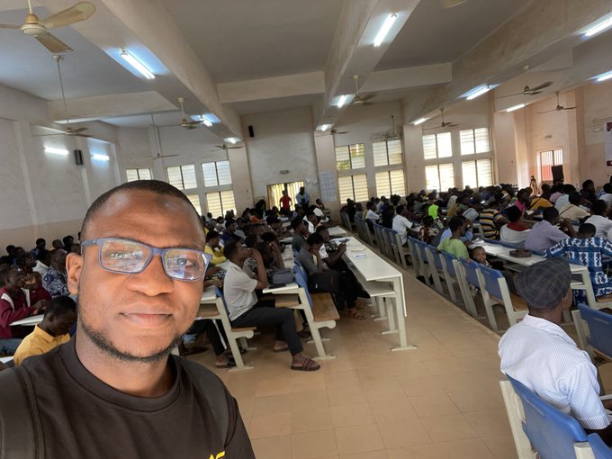 A man takes a selfie in a crowded lecture hall or auditorium filled with students seated at long desks, with no visible text or numbers. The image appears unrelated to the post about a Binance P2P acquaintance and cryptocurrency trading.