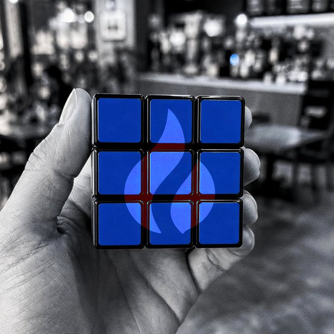 A hand holds a blue Rubik's cube superimposed with a flame logo, set against a blurred black-and-white bar background, symbolizing the energetic start to the workday reflected in the "GMGM! Good morning, world!" greeting.