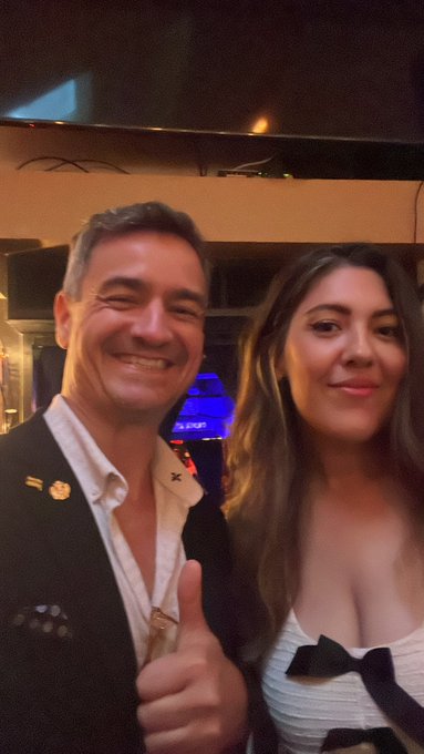A selfie of a smiling man in a dark blazer giving a thumbs up next to a woman with long dark hair wearing a white top with a black bow.