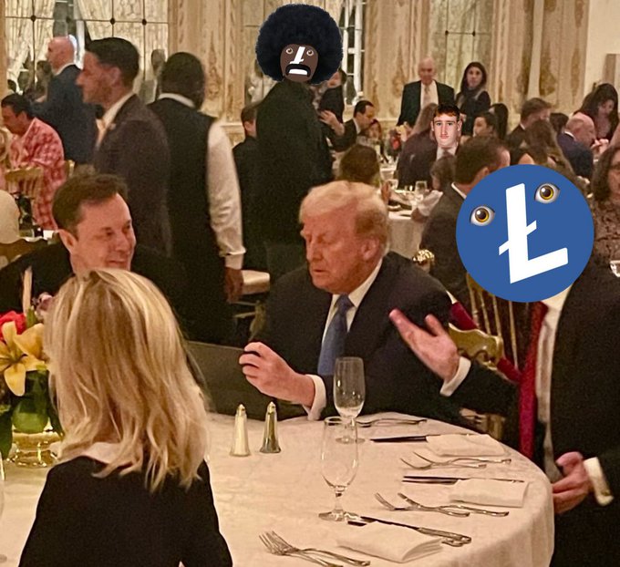 A scene from a formal dinner event shows Donald Trump seated at a table next to Elon Musk, with another person in the background whose face is covered with a digital avatar mask, and a blue 'L' logo emoji added to the image.
